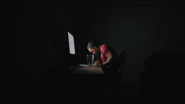 Person in red shirt working at computer desk in dark room with monitor glow