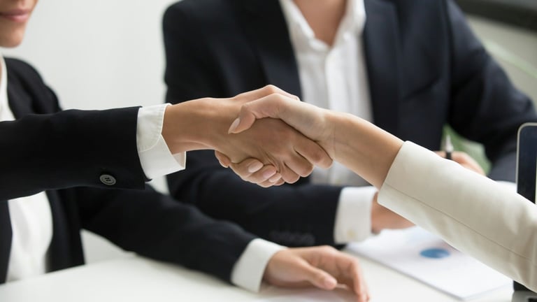Two business professionals in suits shaking hands across a desk during a meeting