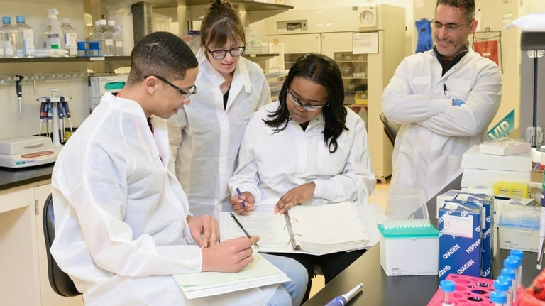 Four scientists in white lab coats working together in a laboratory, reviewing notes and conducting research at a lab bench with equipment.