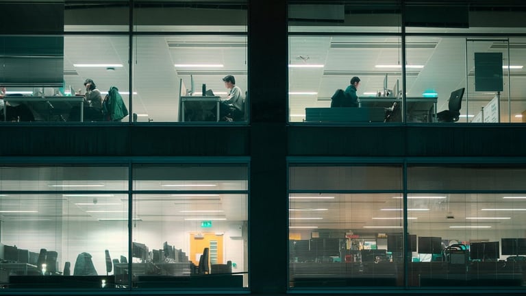 Office building exterior at dusk showing illuminated windows with silhouettes of workers at desks across multiple floors