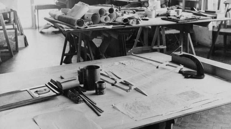 Black and white photo of an architectural workspace with drafting table, tools, blueprints, and construction materials in background.