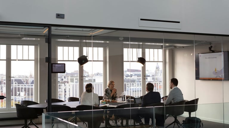 Five people in business meeting around a table in modern office with floor-to-ceiling windows overlooking a city skyline